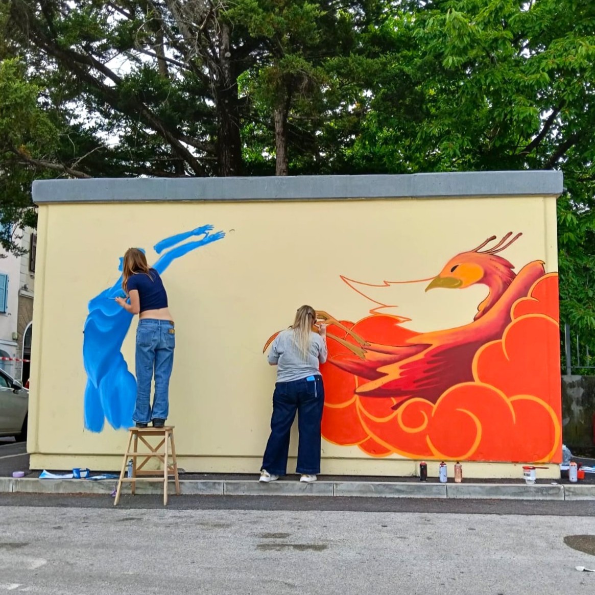 Due ragazze dipingono un murales che raffigura una figura femminile blu a sinistra e una fenice rossa a destra